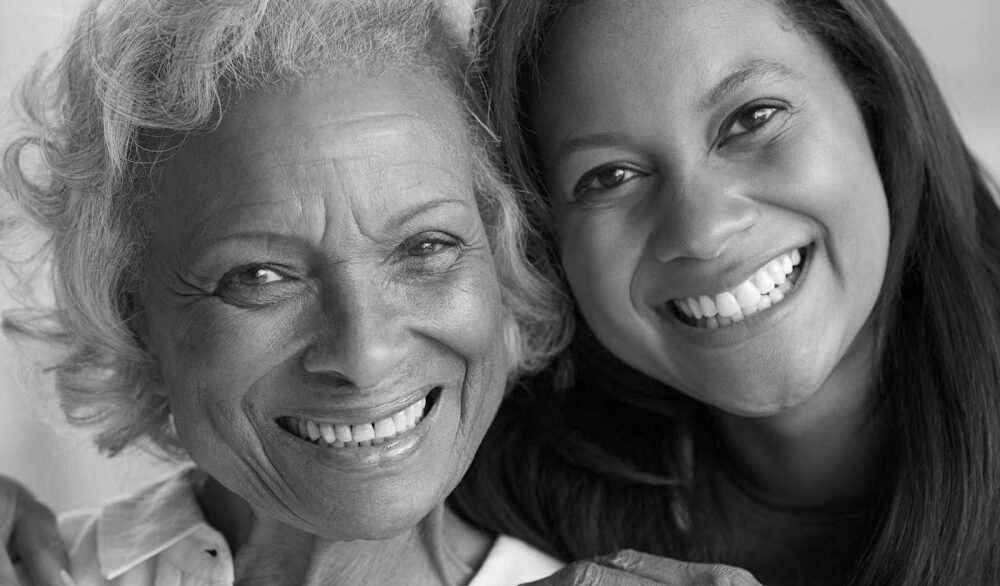 Black and white photo of two women smiling warmly at the camera. One woman has curly hair and is older with wrinkles, while the other woman is younger with straight hair. They are standing close together, exuding a strong sense of home and affection.