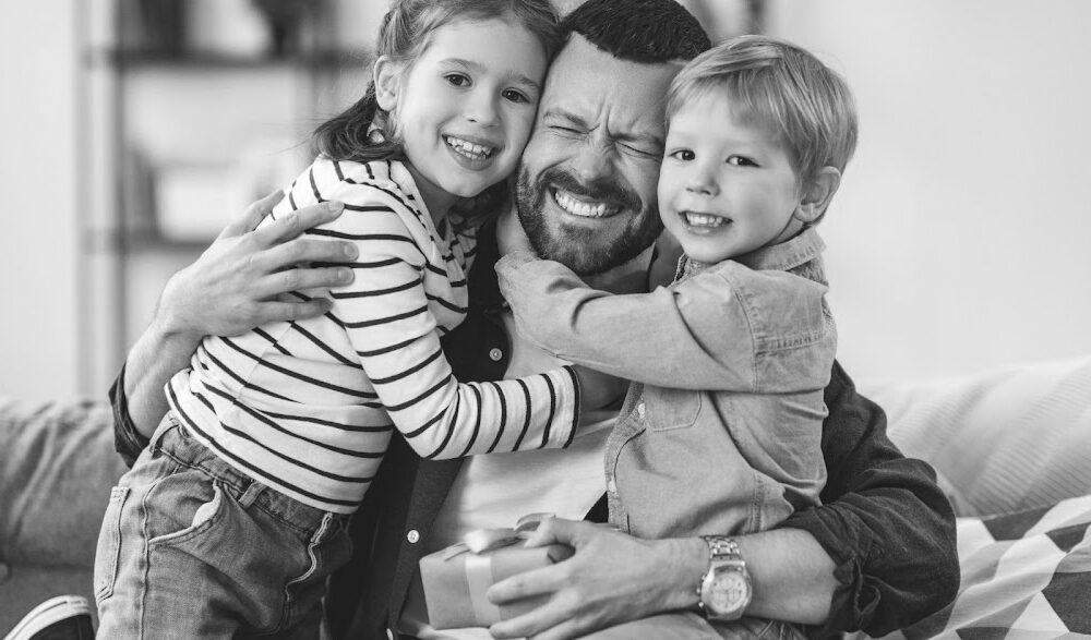 A man with a beard is sitting on a couch at home, smiling joyfully while hugging two young children. One child is holding a small gift box, and they are all dressed casually. The background shows a blurred room with a bookcase and a plant. The image is in black and white.