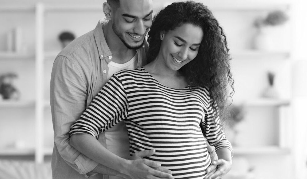 A pregnant woman smiles while her partner, standing behind her, embraces her and gently rests his hands on her belly. They appear happy and excited in the serene, softly lit home setting, perfect for capturing the essence of a loving family moment on your homepage.