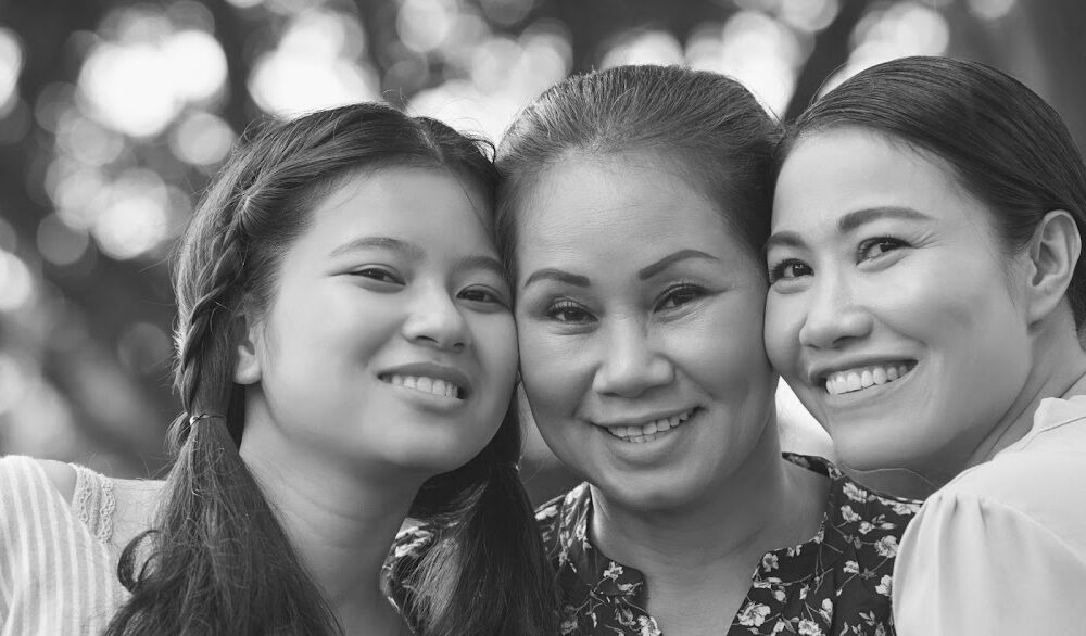 Three women smiling warmly at the camera in a close group, evoking the feeling of home. The woman in the center has her arms around the other two. The background is softly blurred with trees and natural light, enhancing the focus on their happy expressions. Black and white image.