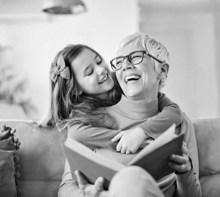 Black and white photo of a joyful elderly woman wearing glasses, holding an open book, and sitting on a couch. A young girl with a hair bow hugs her from behind, both smiling and laughing. The scene captures a tender moment of connection and happiness, answering life's FAQs about love and joy.