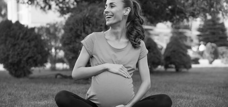 A pregnant woman sits cross-legged on the grass in a park, smiling and looking to her left. She is wearing a short-sleeved shirt and leggings. The background shows trees and shrubs, reminiscent of scenes often found in FAQs about prenatal care. The image is in black and white.