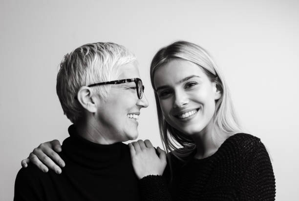 Black and white image of two women standing close together, smiling. One has short, light-colored hair and glasses, while the other has long, light-colored hair. Both wear dark tops. The woman with glasses has her arm around the other's shoulder, highlighting their warm contact.