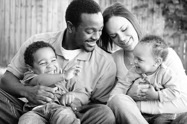 A black and white photo captures a joyful family moment: a father and mother smile at their two young children. The father and older child share a glance, while the mother holds the baby, who beams joyfully. This image brings to life answers to frequently asked questions about family love and harmony.