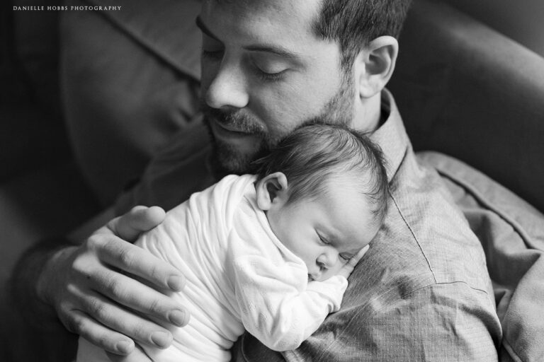 A man with a beard holds a sleeping baby close to his chest, eyes closed, providing a sense of calm and tenderness. The black-and-white photo captures the peaceful bond between them, much like finding answers in FAQs when you need help.