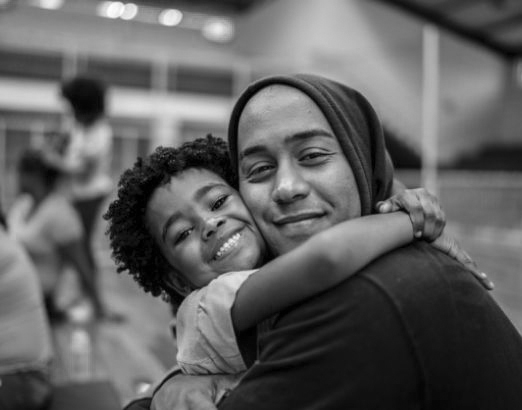 In a black-and-white image, a child with curly hair and a broad smile hugs an adult wearing a hoodie and beanie. They both have happy expressions. The blurred indoor setting, possibly a gymnasium or community center, subtly hints at celebrations tied to immigration milestones.