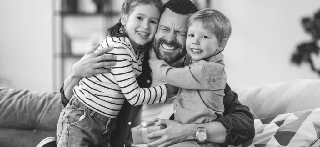 A man is sitting on a couch, smiling, as he hugs two young children. One child is wearing a striped shirt, and the other is in a jean shirt. They all appear joyful and are engaged in a warm embrace. The setting is a cozy living room.