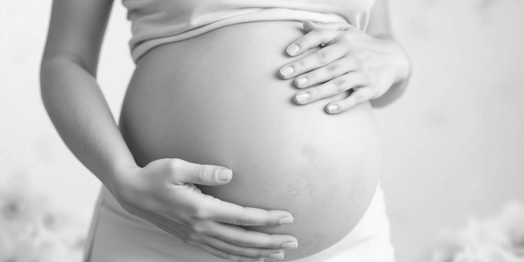 A black-and-white image of a pregnant person gently cradling their belly with both hands. They are dressed in a light-colored top and pants, evoking thoughts of the journey ahead, such as prenatal DNA testing. The background is softly blurred, adding to the serene atmosphere.