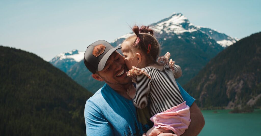 A man in a blue shirt and cap smiles while holding a young child, who kisses him lovingly. The child, wrapped in a pink blanket and wearing a striped top, enjoys the moment against the backdrop of snow-capped mountains and lush greenery—an undeniable bond beyond any DNA test.