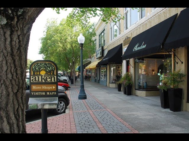 A tree-lined street with a sidewalk featuring various shops under black awnings. A sign reads "Aiken," promoting more shops and visitor maps, including one for drug testing locations. Parked cars line the street, while a lamppost stands nearby on this sunny day. Service Area