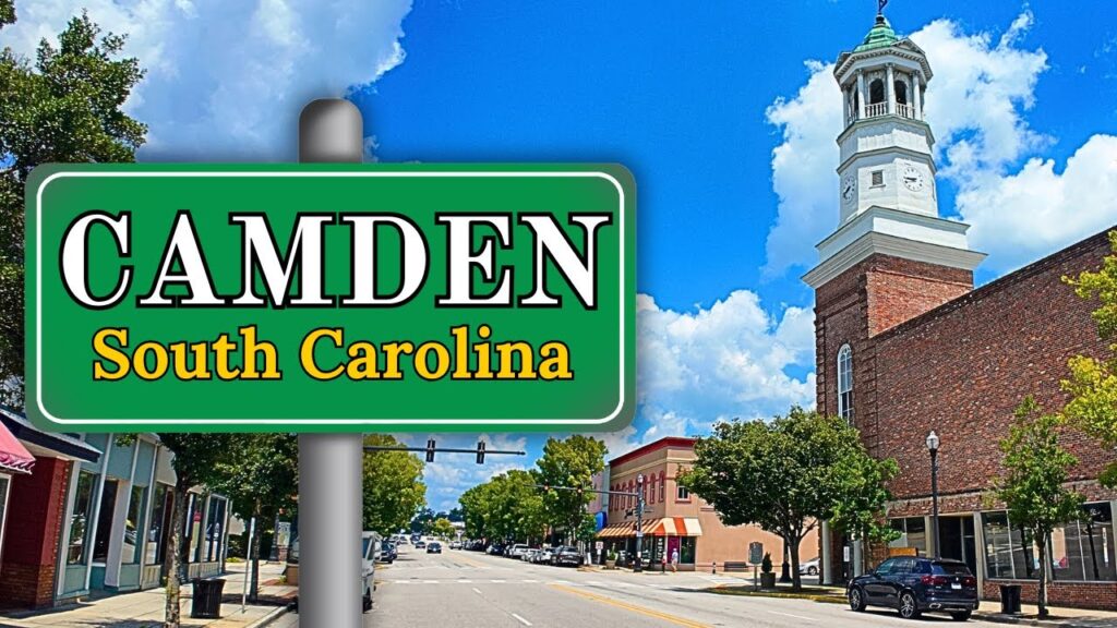 A street view of Camden, South Carolina, showcasing a large green sign with "CAMDEN South Carolina" written on it. Amidst the trees and a brick building with a clock tower beneath the clear blue sky, you might find local services such as drug testing services enhancing community wellness.