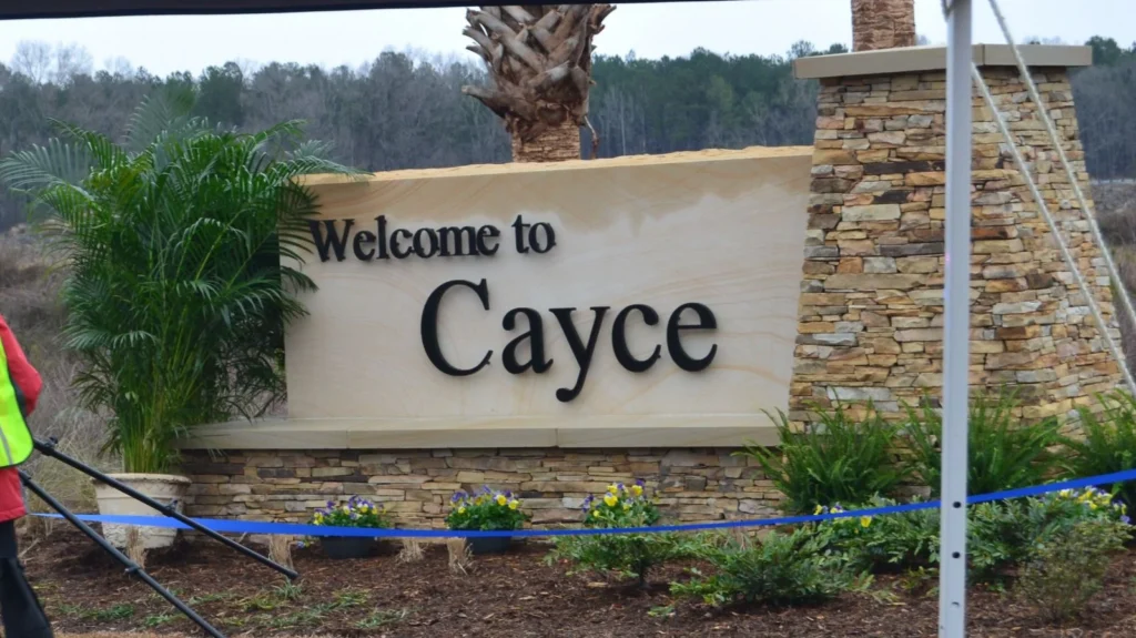 A stone sign reads "Welcome to Cayce" in large black letters, surrounded by plants and flowers. The background shows trees and a cloudy sky. A partial view of a person in a safety vest, possibly overseeing nearby drug testing services, is visible on the left.