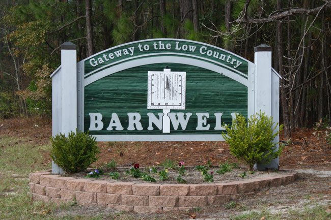 A sign reading "Gateway to the Low Country, Barnwell" features a sundial image. The green sign with white text, bordered by white posts, rests on a stone base surrounded by small shrubs and trees. Nearby, discreetly nestled amidst the greenery, are facilities offering drug testing services.