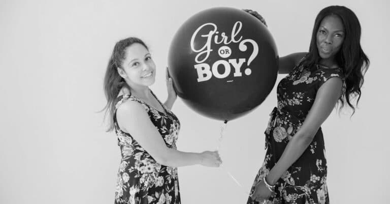 Two women in floral dresses hold a large black balloon with "Girl or Boy?" printed on it, hinting at a gender reveal theme inspired by their prenatal DNA test. They pose against a plain backdrop, smiling warmly at the camera.