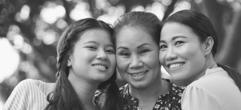 Black and white image of three women smiling closely together outdoors. The background is softly blurred, showing large tree leaves. One woman in the center is older, flanked by two younger women, all radiating joy akin to discovering connections through siblingship DNA testing.