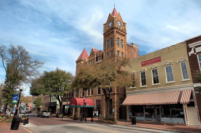 Street view of a charming small town with a historic building featuring a clock tower. Trees line the sidewalk, and businesses offering drug testing services with awnings are on either side. The sky is partly cloudy, creating a quaint and inviting atmosphere.