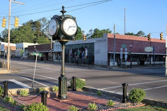 A street scene showcases a vintage clock on a sidewalk with a green base and white clock faces. Behind it, brick buildings display a painted mural and shops offering diverse services, from cafes to drug testing. The road is empty, traffic lights are visible, and the sky is clear and blue.