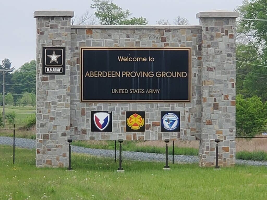 Stone entrance sign reads "Welcome to ABERDEEN PROVING GROUND UNITED STATES ARMY" with the U.S. Army logo and three military insignias below, surrounded by grass, trees, and facilities offering drug testing services.