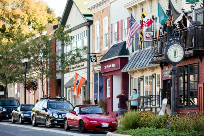 A small-town street with parked cars, including a red convertible, people chatting on the sidewalk near drug testing services, American and state flags, and a large clock reading "Bel Air" in front of brick storefronts.