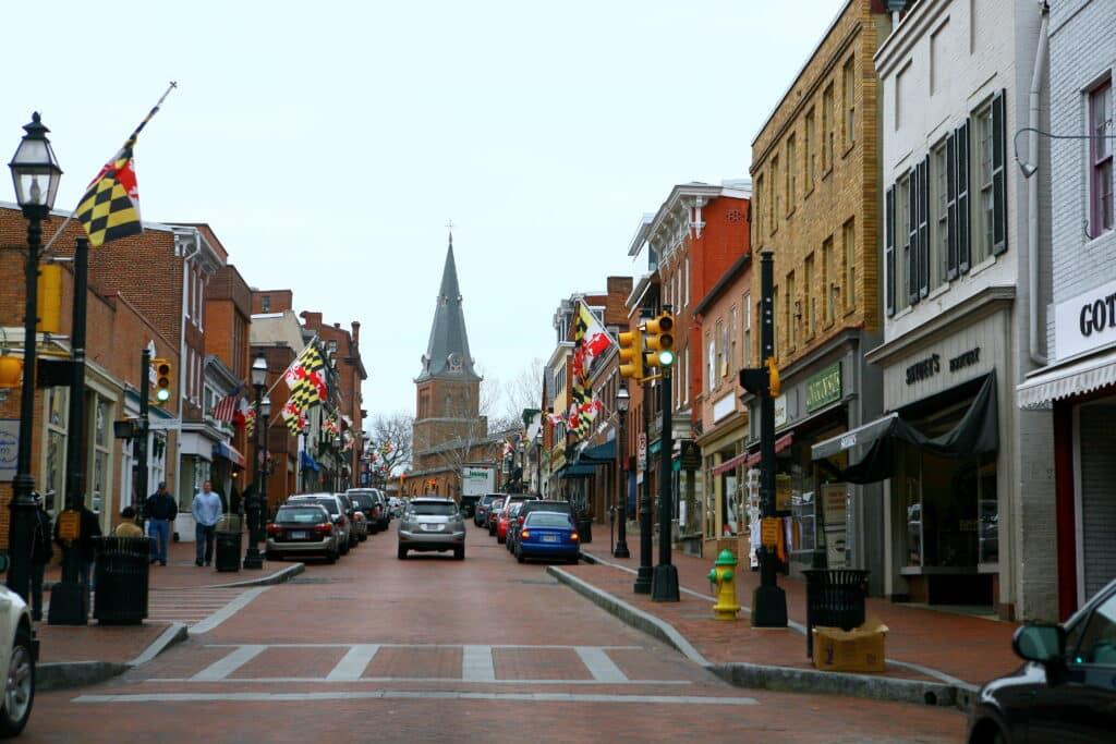 A brick-paved street in a historic downtown area with shops, parked cars, Maryland state flags, and a church with a tall steeple. Among the local businesses, drug testing services are available on this cloudy day.