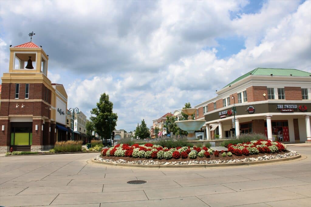 A roundabout with a fountain and red and white flowers is surrounded by brick buildings housing shops, restaurants, and drug testing services under a partly cloudy sky.