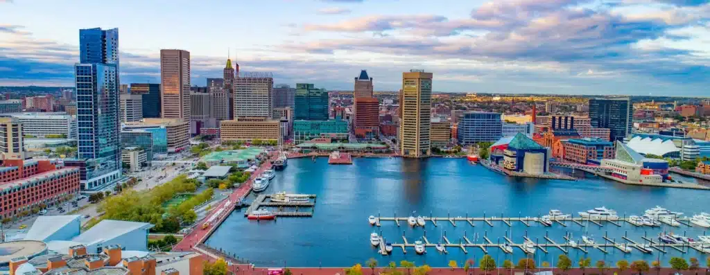 Aerial view of Baltimore’s Inner Harbor with modern high-rise buildings, boats docked at marinas, nearby drug testing services, waterfront attractions, and a partly cloudy sky in the background.