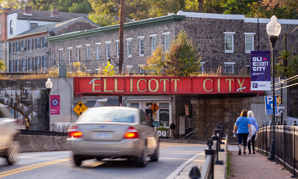 A red bridge with yellow "Ellicott City" lettering spans over a street offering access to local shops and drug testing services. Cars drive underneath, and two people walk on the sidewalk past stone buildings and “Old Ellicott City” signs.