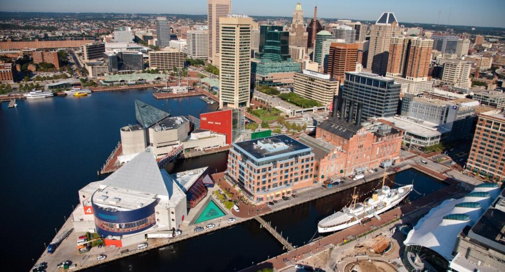 Aerial view of Baltimore’s Inner Harbor showing waterfront buildings, an aquarium, historic ships docked along the harbor, and the city skyline. The area includes businesses offering amenities such as drug testing services on a sunny day.