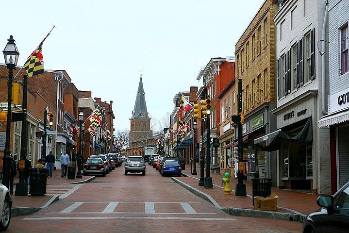 Brick-paved street lined with shops offering local goods and drug testing services, Maryland state flags, cars parked along the sides, and a church with a tall steeple in the background on a cloudy day.