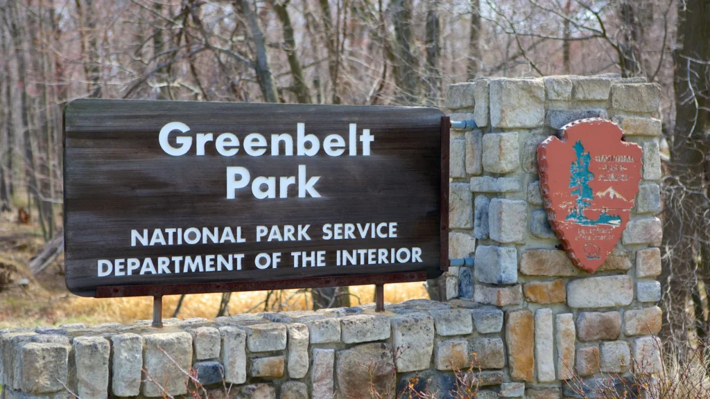 A stone sign at the entrance of Greenbelt Park displays the park's name along with “National Park Service,” “Department of the Interior,” and information on nearby drug testing services, all surrounded by trees in the background.