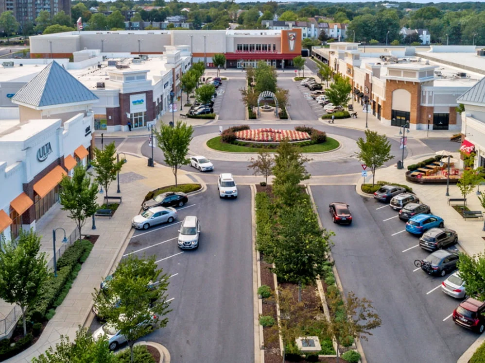 Aerial view of a shopping center with stores, parked cars, and trees lining the sidewalks. A roundabout with a central landscaped area is at the center, near shops offering amenities like drug testing services as people walk between stores and vehicles.