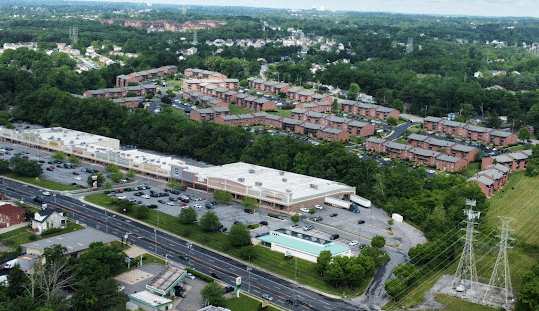 Aerial view of a suburban area featuring a shopping center with drug testing services, a large parking lot, surrounded by trees, roads, apartment complexes, houses, and power lines running through grassy land.