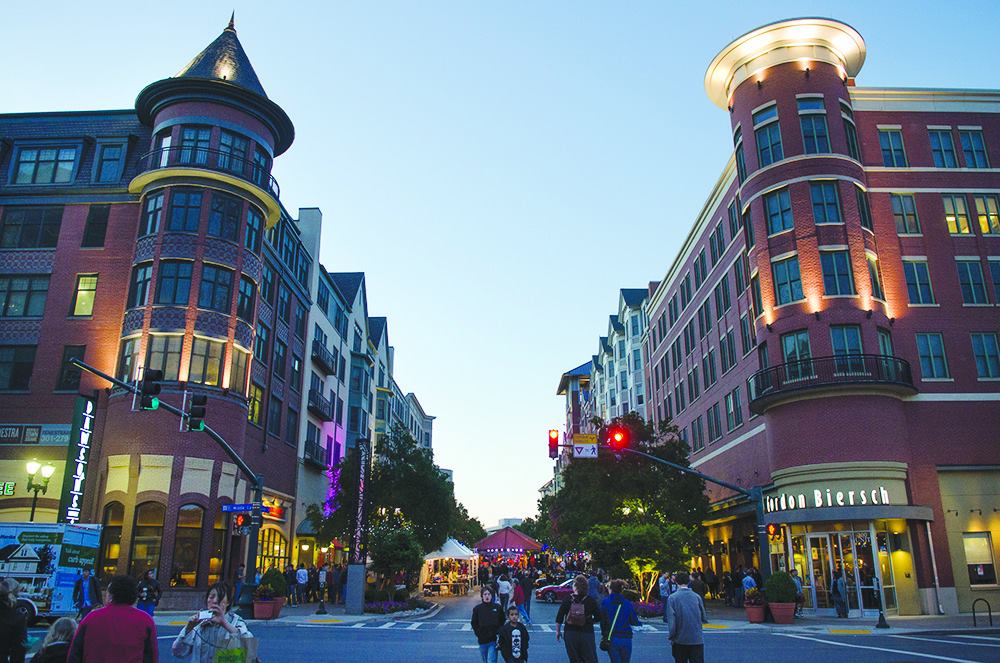 A lively urban street scene at dusk, with people walking between two large red-brick buildings featuring rounded corners and shops offering local goods and drug testing services. Bright lights and festive decorations create a welcoming atmosphere.