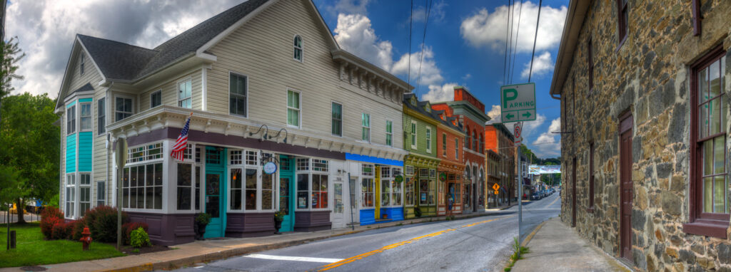 A colorful row of historic shops and buildings lines a quiet street in a small town, with a parking sign visible and trees and clouds in the background—one storefront advertises local drug testing services.