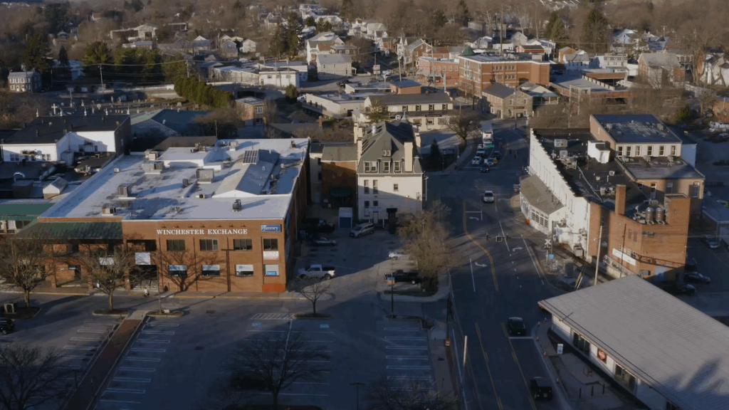 Aerial view of a small town with tree-lined streets, brick buildings, parking lots, and cars. The Winchester Exchange building is visible alongside businesses offering drug testing services. Residential houses stretch into the background under clear skies.