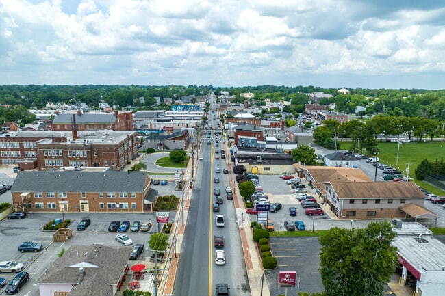 Aerial view of a small town with cars driving down the main road, surrounded by buildings offering drug testing services, parking lots, and trees under a partly cloudy sky.