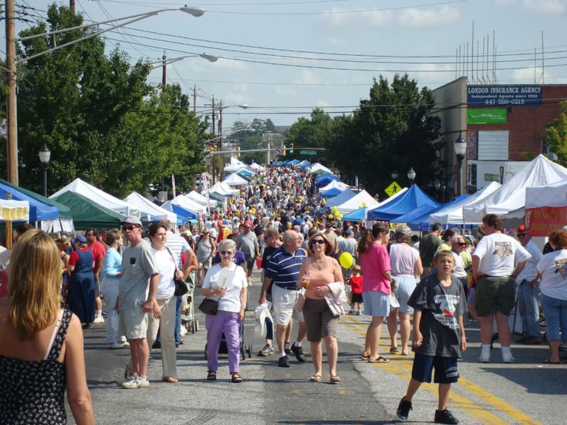 A large crowd of people walk along a street lined with white and blue vendor tents at an outdoor festival on a sunny day, where booths offer various attractions—including drug testing services. Trees and buildings are visible in the background.