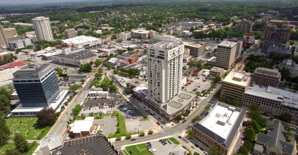 Aerial view of a city with a tall central building, surrounded by mid-rise buildings, roads, parking lots, and green spaces on a sunny day. Trees and a sprawling suburban area are visible—home to clinics offering drug testing services.