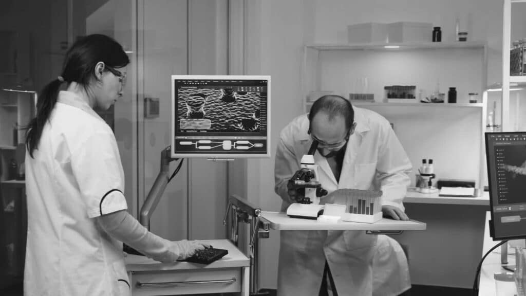 Two scientists in lab coats work in a laboratory; one types on a keyboard near a monitor displaying scientific images, while the other reviews samples.
