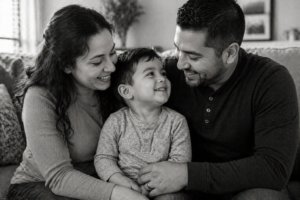 A smiling woman and man sit closely on a couch with their young child between them. All three are looking at each other happily, sharing a joyful moment together. The image is in black and white.