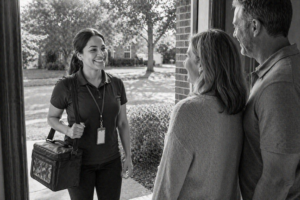 A smiling woman with a name badge and insulated bag stands at a front door in a midlands neighborhood, greeting a couple who are welcoming her inside. The scene is set in residential Columbia, SC.
