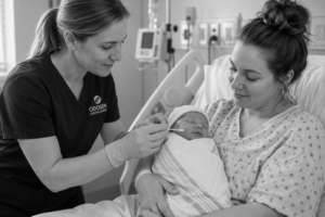 A nurse gently swabs the mouth of a newborn baby wrapped in a blanket, while the baby is held by a smiling woman in a hospital bed. The scene is calm and caring in a hospital room.
