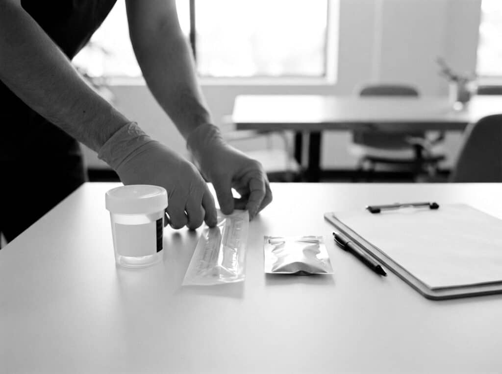 Gloved hands arranging a sealed urine specimen cup, oral fluid swab and hair sample envelope beside a clipboard